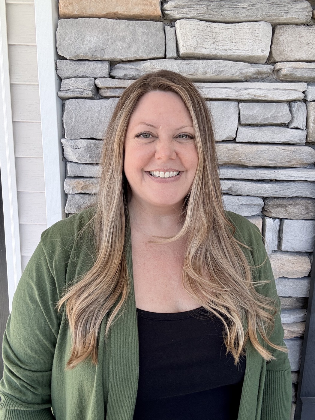 Nicole Folker smiling, wearing a green cardigan, in front of a stone wall